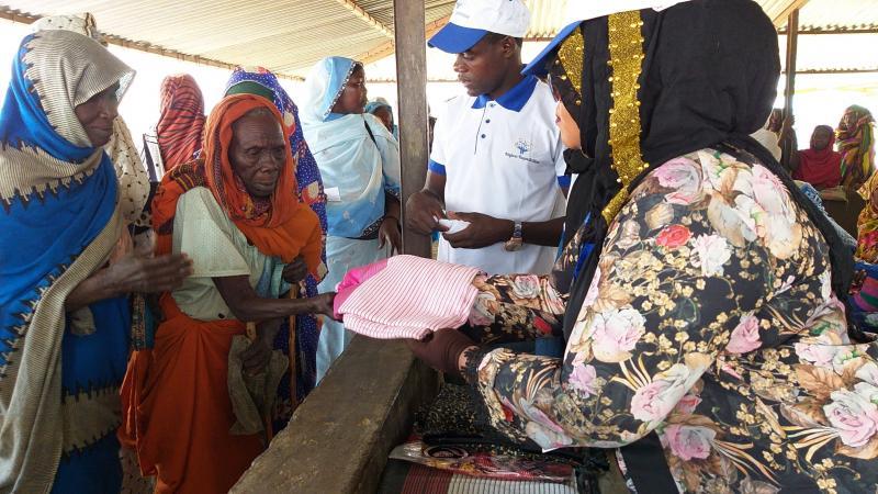 Najwa Adam donating new clothing to a group of women refugees in Africa. The women is reaching her hand out to receive the pink piece of clothing.
